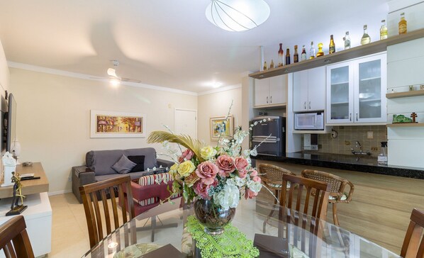 A dining area with a glass table, chairs, and a vase of flowers. There is a sofa and a television in the background.