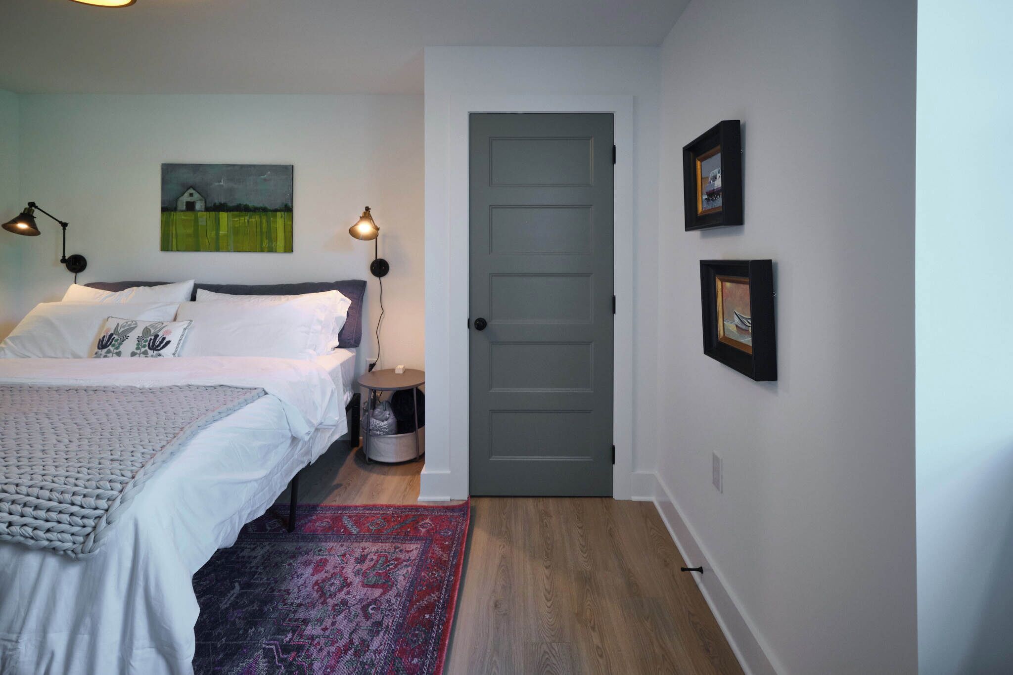 A bedroom with a bed, bedside lamps, a rug, a door, and wall art.