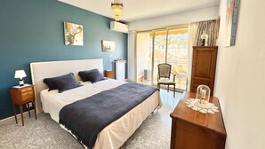 A bedroom with a large bed, a chandelier, a view through a sliding glass door, and a wooden nightstand with a lamp. - 100M FROM THE CASINO, LOVELY 3-ROOM APARTMENT WITH SEA VIEW AND BIJOU GARDENS. PRIVATE PARKING. (Menton)