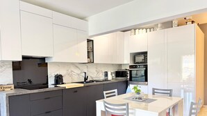 A modern kitchen with white cabinets, a marble backsplash, a dining area with a white table and red chairs, and a small island with a sink. - 100M FROM THE CASINO, LOVELY 3-ROOM APARTMENT WITH SEA VIEW AND BIJOU GARDENS. PRIVATE PARKING. (Menton)
