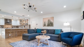 Un salon moderne avec un canapé d’angle, une table basse ronde et un fauteuil bleu. On aperçoit une cuisine avec un bar au fond.