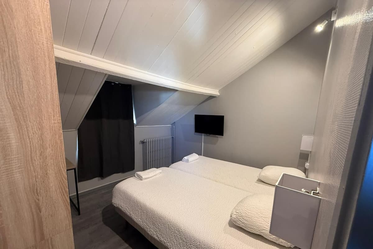 A modern bedroom with a sloped ceiling, a bed with a white comforter, a flat-screen TV mounted on the wall, and a radiator.