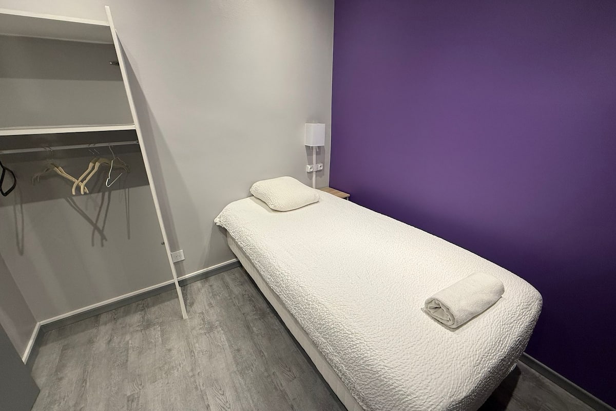 A small, single-bed room with a grey wardrobe, a purple wall, and a wooden floor.