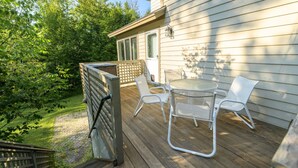 Una terraza de madera con mobiliario exterior, un vidrio que da a un espacio cubierto y vista a un jardín exuberante.