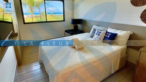 Une chambre à coucher avec un lit, des oreillers, une table de chevet, une lampe et une vue sur la plage par la fenêtre.