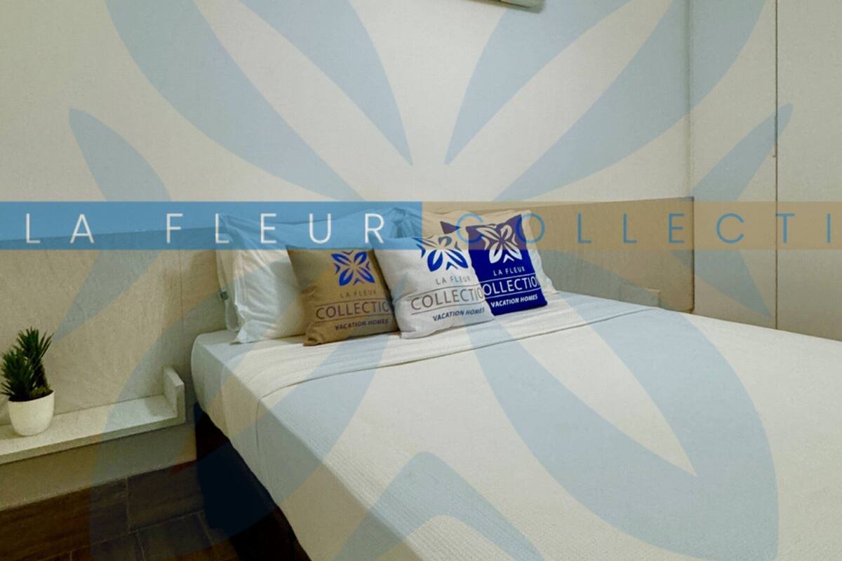 A bedroom with a bed, pillows, a small bedside table, a potted plant, and an air conditioning unit on the wall.