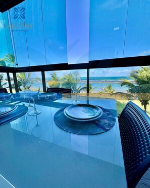 A glass-top dining table with plates and wine glasses, overlooking a beach with palm trees and the ocean.