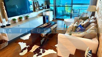 A modern living room with a flat-screen TV, a sofa, a coffee table, and a large window offering a view of the ocean.