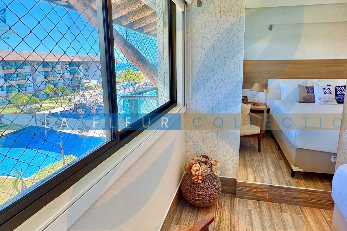 Ein Hotelzimmer mit großem Fenster, das einen Blick auf einen Pool und Gebäude bietet, ein Bett mit weißer Bettwäsche und ein kleiner dekorativer Korb auf dem Boden.