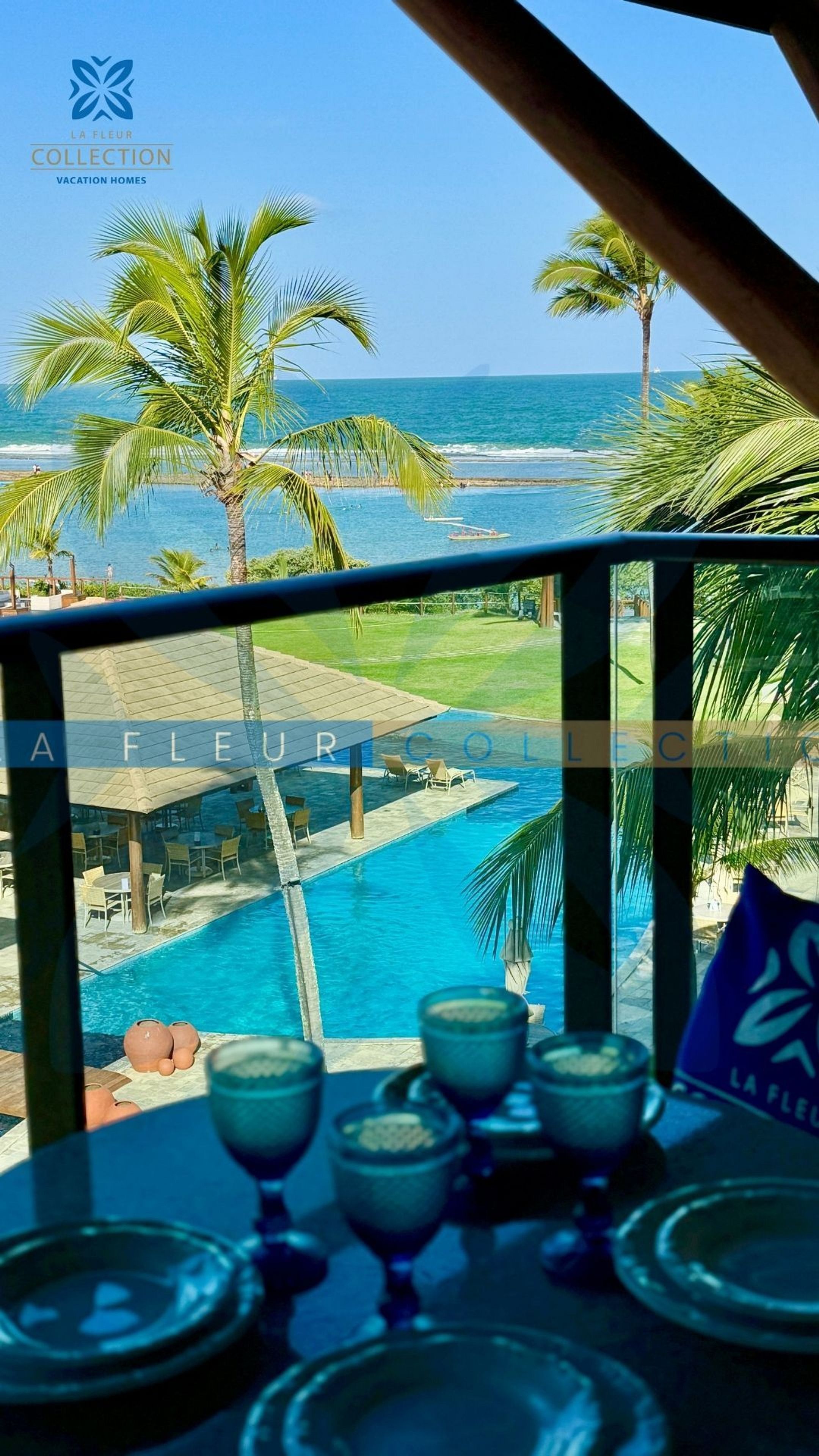 A poolside view with a clear view of the ocean, palm trees, and a lounge area with tables and chairs.