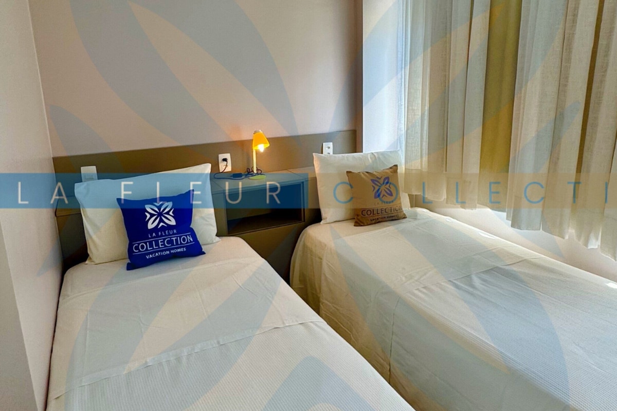 A hotel room with two beds, a nightstand, a lamp, and a window with curtains.