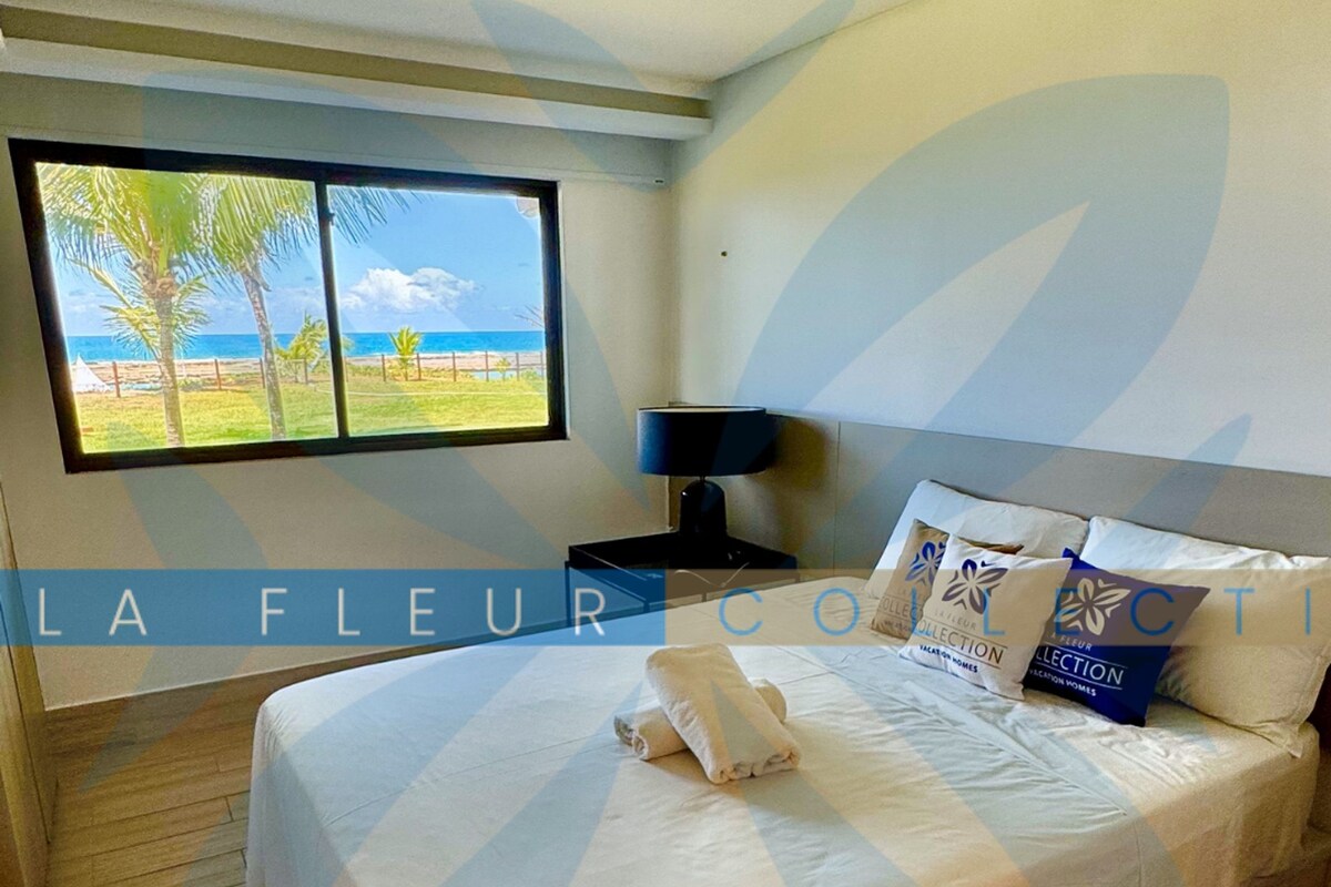 A bedroom with a large bed, a nightstand, a lamp, and a window with a view of the beach.