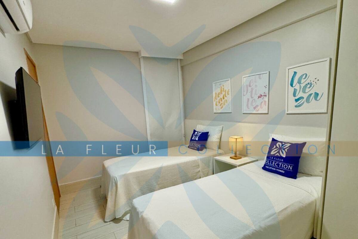 A hotel room with two beds, a small table, a wall-mounted TV, and a ceiling light.