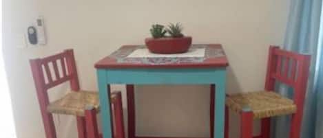 Una habitación pequeña con una mesa y dos sillas, una planta en maceta sobre la mesa, y una ventana con cortinas.
