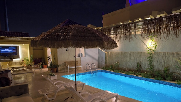 A swimming pool with lounge chairs and a thatched umbrella, surrounded by plants and a TV screen. - Tayba Alkubra Hostel (Riyadh)