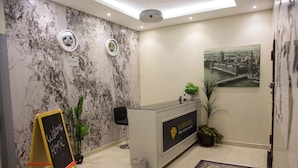 A reception area with a marble wall, a reception desk, a welcome sign, a potted plant, and a cityscape painting. - Tayba Alkubra Hostel (Riyadh)