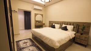 A bedroom with a large bed, a bedside table with a plant, a mirror above the bed, and a door leading to another room. - Tayba Alkubra Hostel (Riyadh)