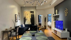 A modern living room with a dark sofa, a coffee table with green placemats, a TV on a white stand, and a unique ceiling light fixture. - La Galerie Privée Luxe & Impeccable Stay (Montréal)