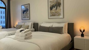 A hotel room with two beds, each with a neatly folded towel, a nightstand with a lamp, and two framed abstract paintings on the wall. - La Galerie Privée Luxe & Impeccable Stay (Montréal)