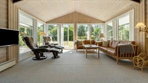 Un salon confortable avec un téléviseur, des fauteuils en cuir, un canapé et une table. De grandes fenêtres offrent une vue sur l’extérieur.