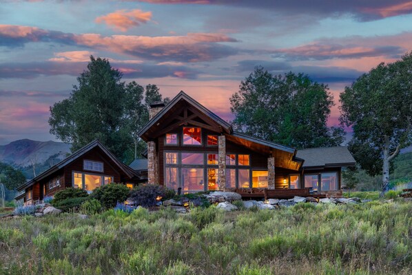 A large, multi-story log cabin with a prominent chimney, surrounded by a natural landscape with trees and a mountainous backdrop. - Telluride Modern Estate | Panoramic Mountain Views (Telluride)
