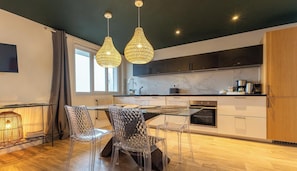 A modern kitchen with a dining area, featuring a glass table, clear chairs, and pendant lighting.