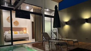 A modern room with a bed, a lounge area with chairs, a small pool with green lighting, and a view of the outdoors.