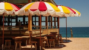 An outdoor seating area with striped umbrellas, wooden tables, and chairs, overlooking the sea.