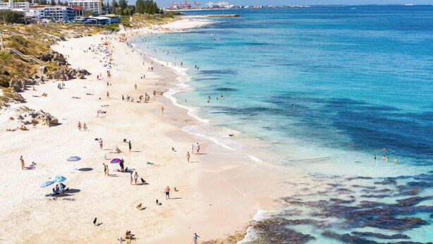 A beach with clear turquoise water, sandy shore, and a few buildings in the background.