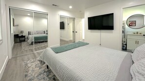 Un dormitorio con una cama grande, un televisor montado en la pared, un espejo sobre el lavabo y un vestidor.
