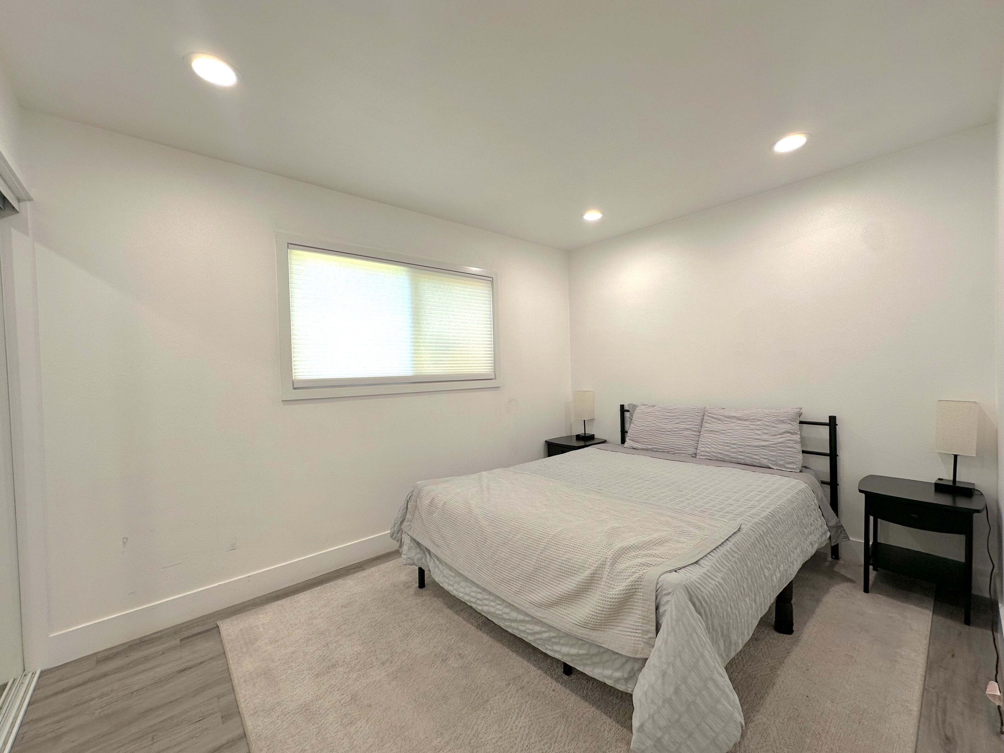 A bedroom with a bed, two bedside tables, a window, and recessed ceiling lights.
