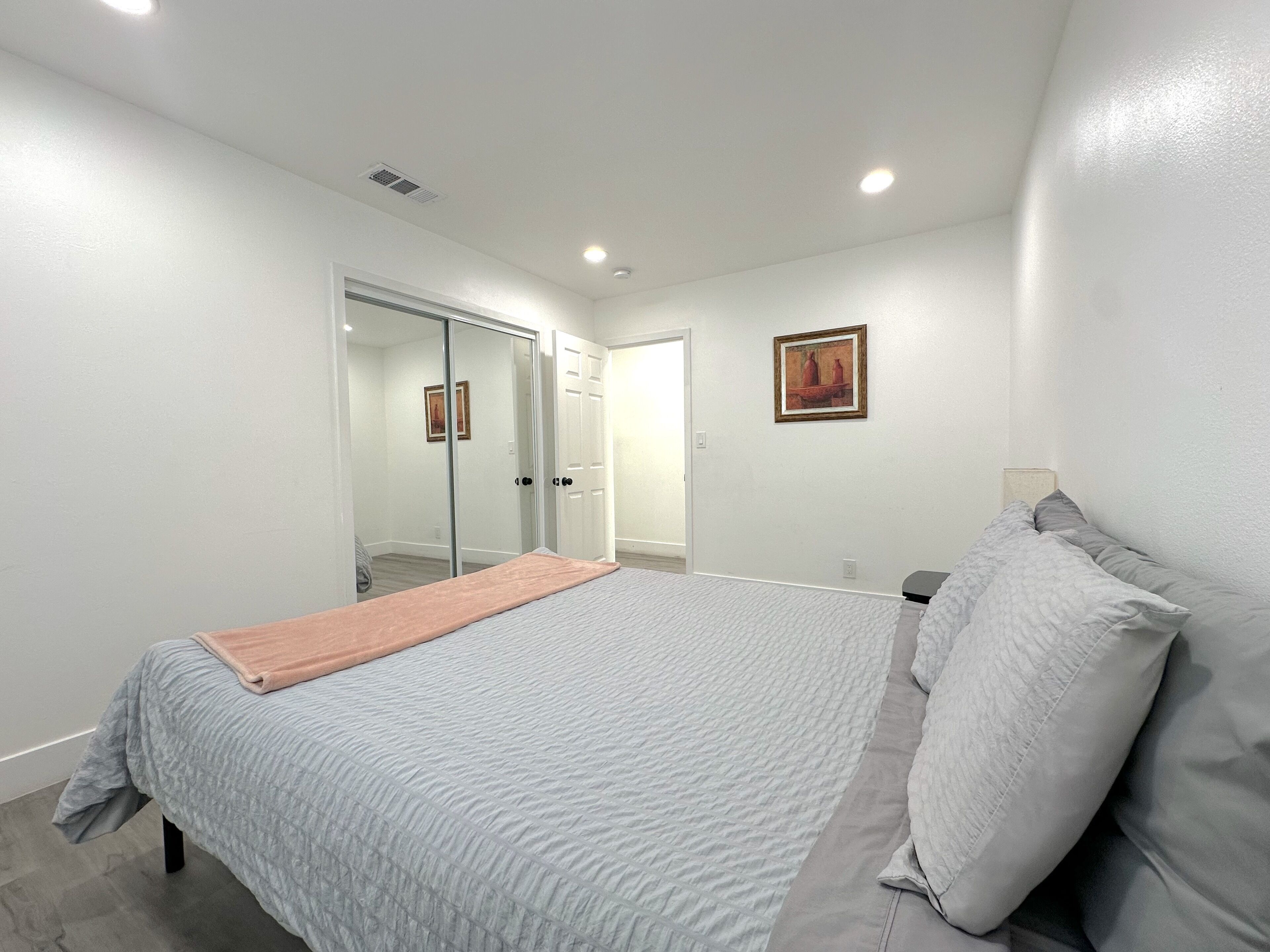Une chambre à coucher comprenant un lit, une table de chevet, une commode, un tableau accroché au mur et une porte-fenêtre coulissante donnant sur une autre pièce.