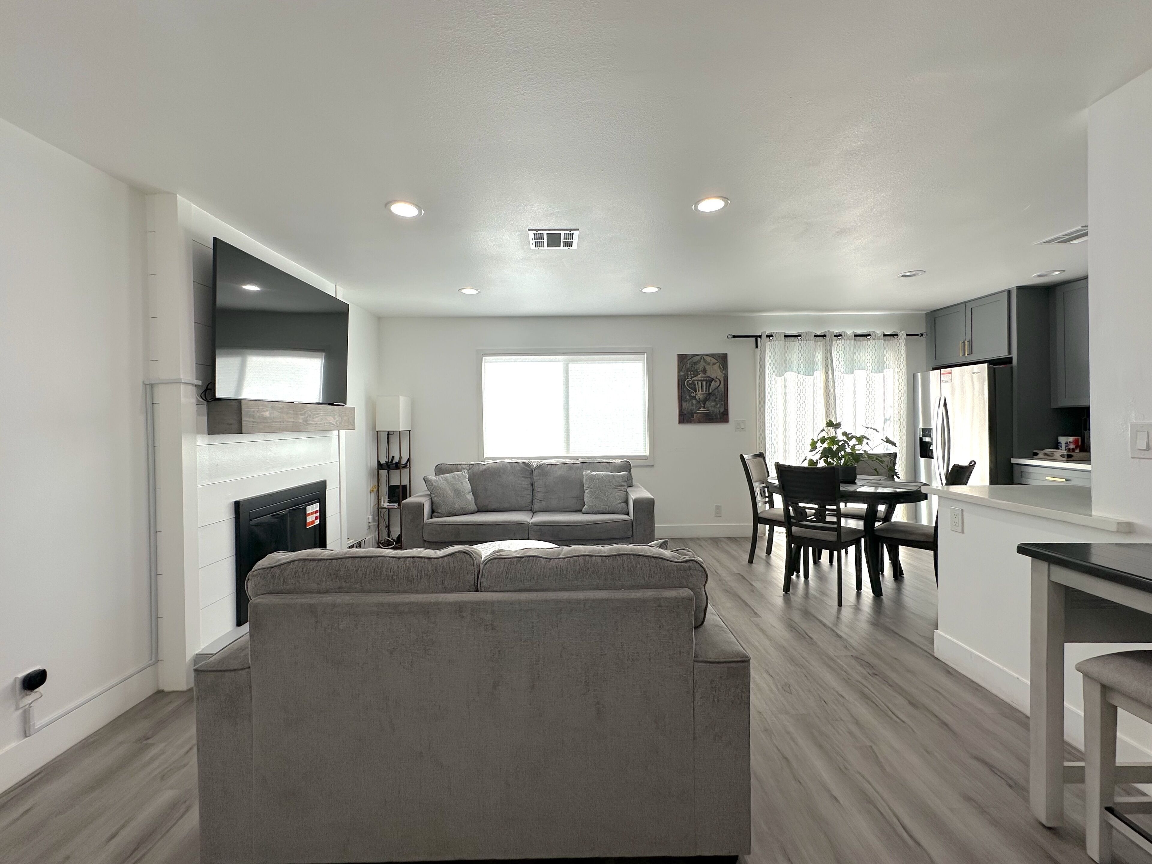 Una sala de estar moderna con un sofá gris, un comedor con una mesa y sillas, y una cocina con gabinetes grises y un refrigerador blanco.