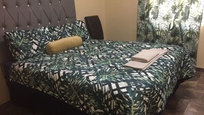 A bed with a tropical leaf pattern bedspread, a tufted headboard, and a dark-colored chair. - Chobe Oasis Guest House (Kasane)