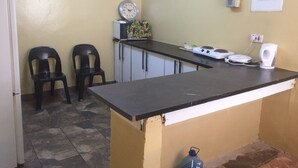 A compact kitchen with a stove, a kettle, and a clock on the wall. There are two chairs and a refrigerator. - Chobe Oasis Guest House (Kasane)