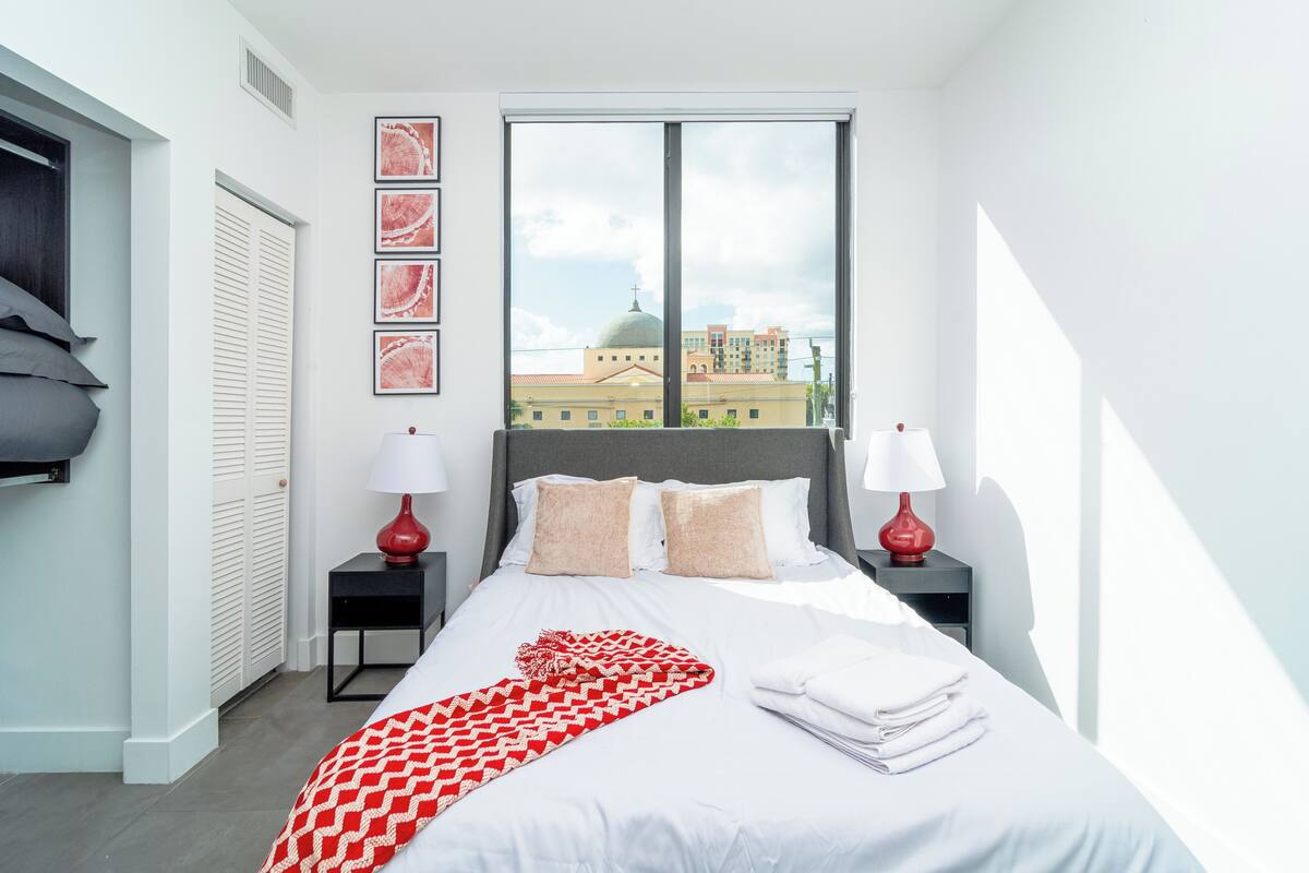 A modern hotel room with a large bed, two bedside tables with lamps, a view of a building with a dome through a large window, and a red and white patterned throw blanket on the bed.