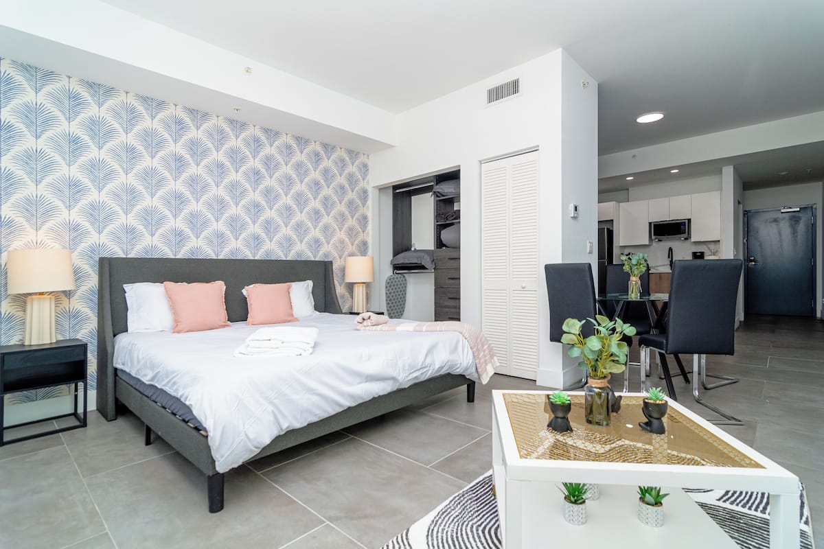 A modern bedroom with a large bed, bedside tables, a coffee table with plants, and a view into the kitchen area.