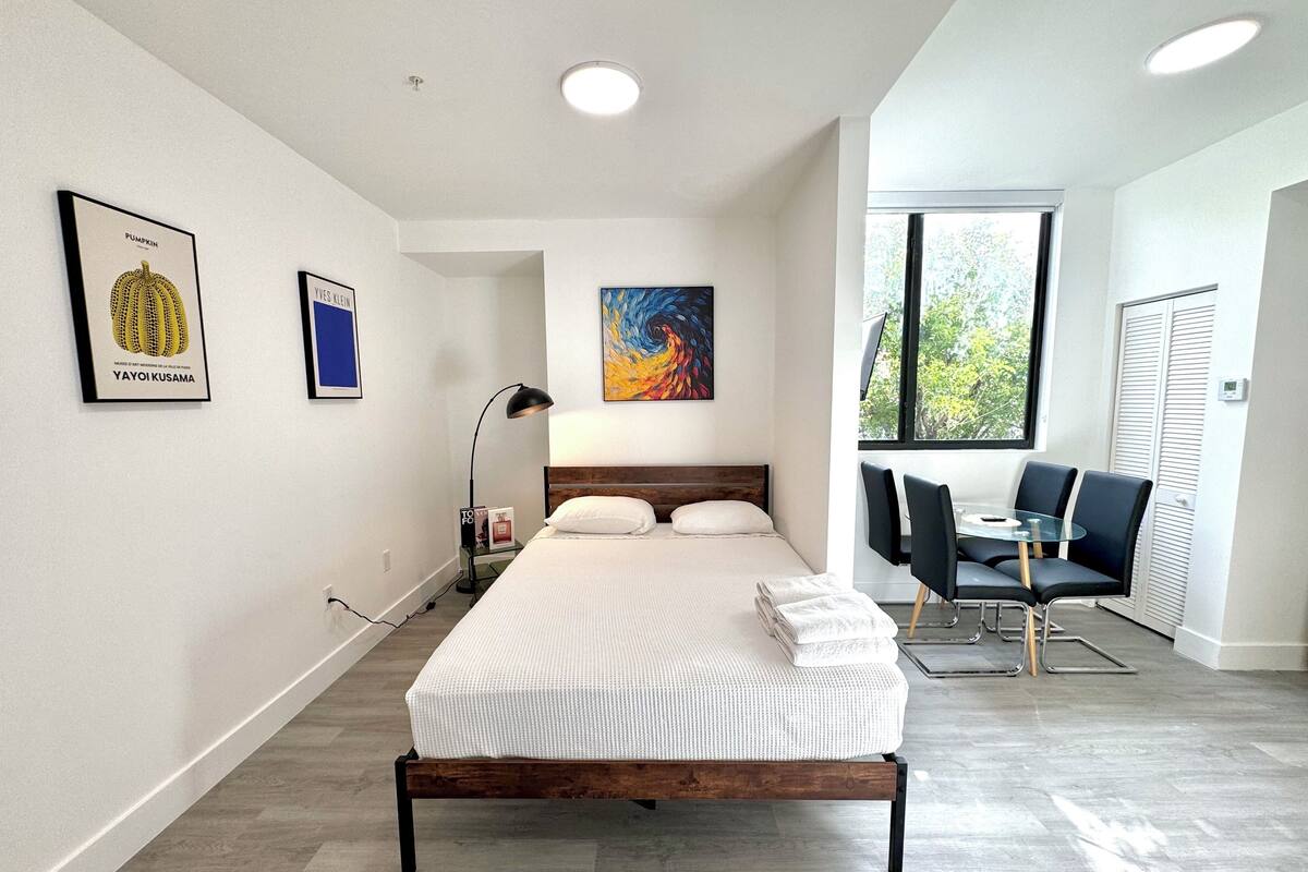 A modern bedroom with a large bed, a desk, and a chair. There are framed pictures on the wall and a window with a view of trees.