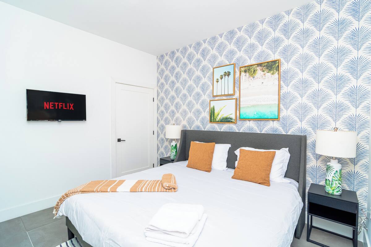A neatly made bed with a striped blanket, a wall-mounted TV labeled 'NETFLIX', and framed pictures on the wall.