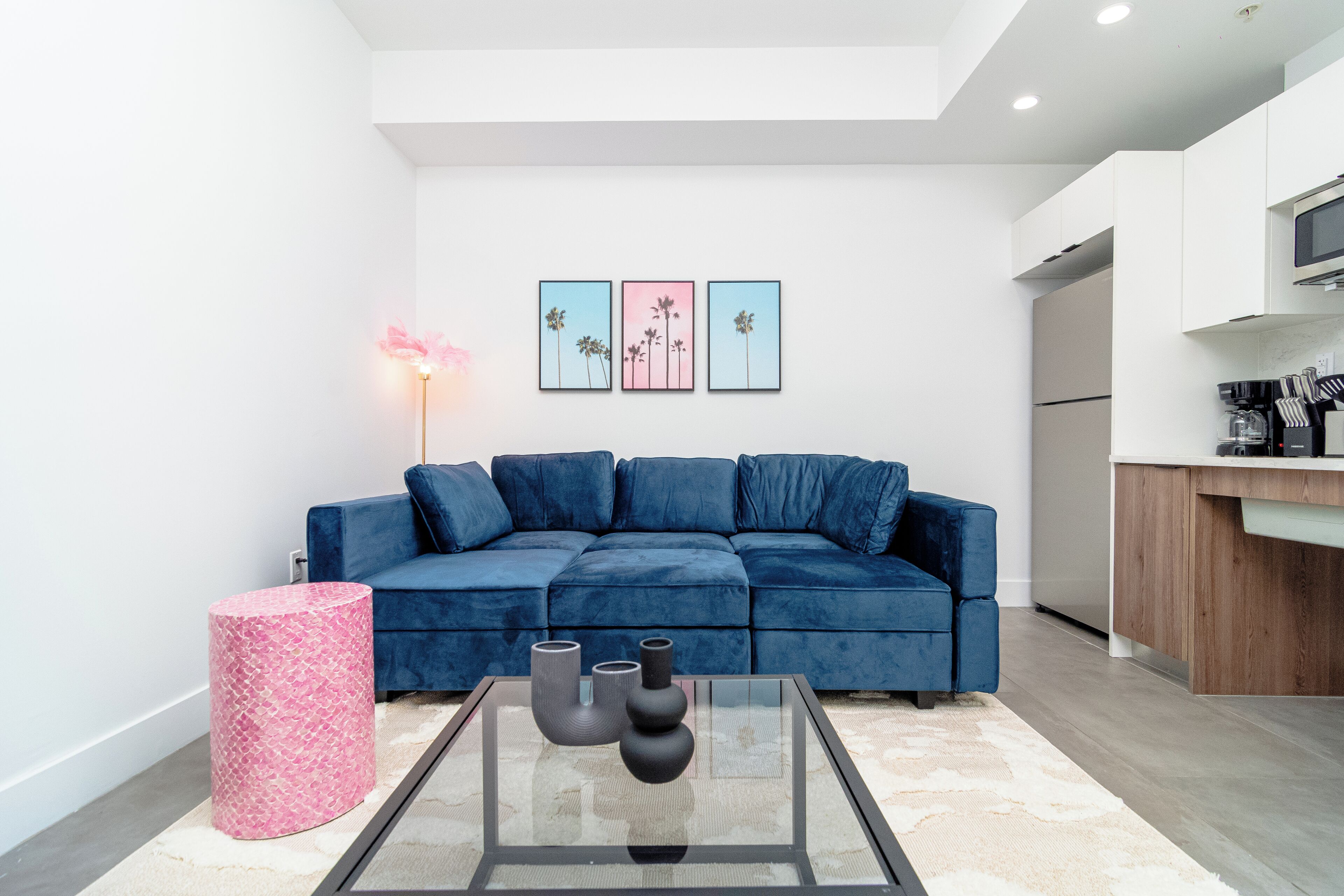 Un salon moderne avec un canapé bleu, une table basse en verre ornée de vases et une petite table d’appoint cylindrique rose.