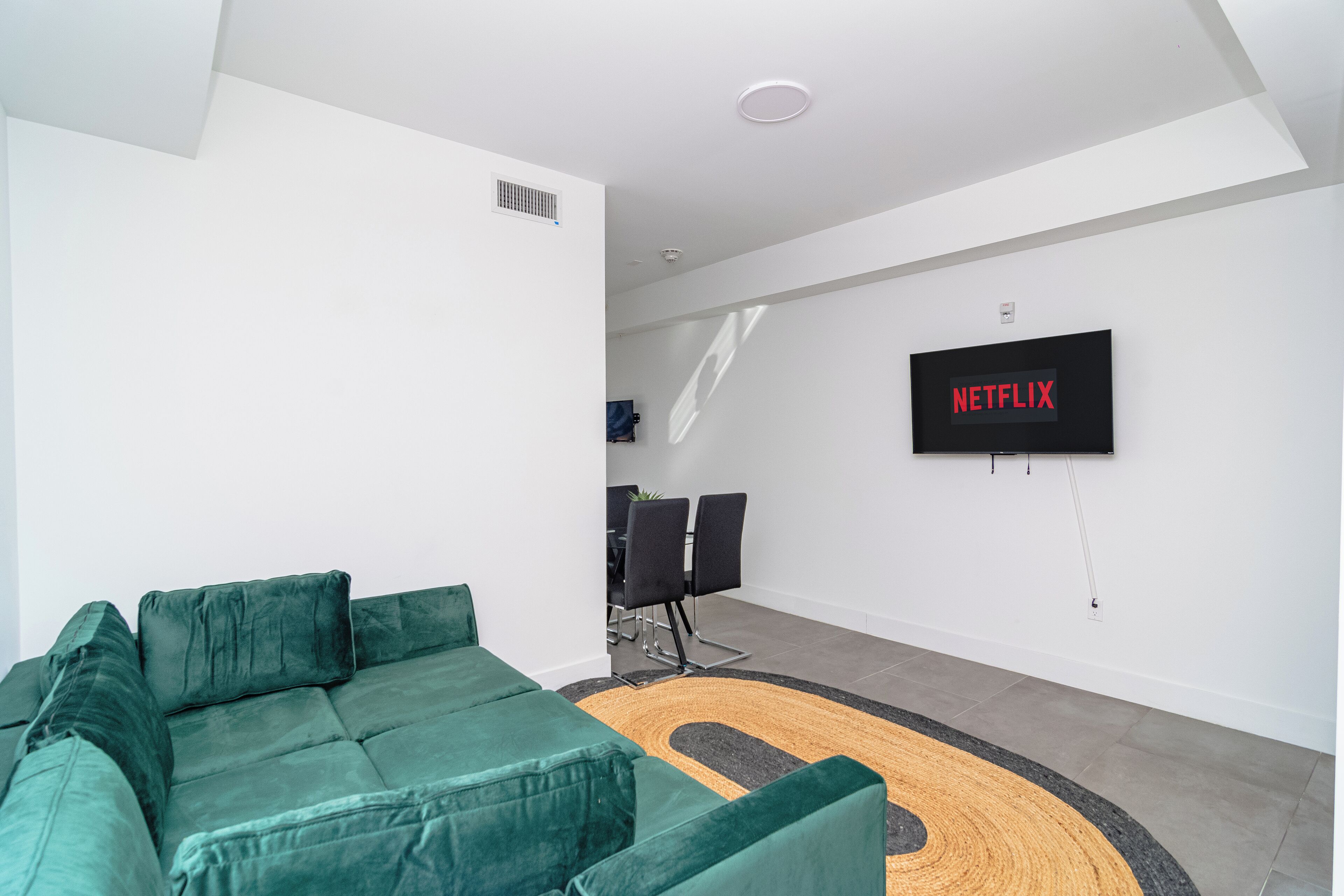 A modern living room with a green sofa, a flat-screen TV mounted on the wall, and a dining area in the background.