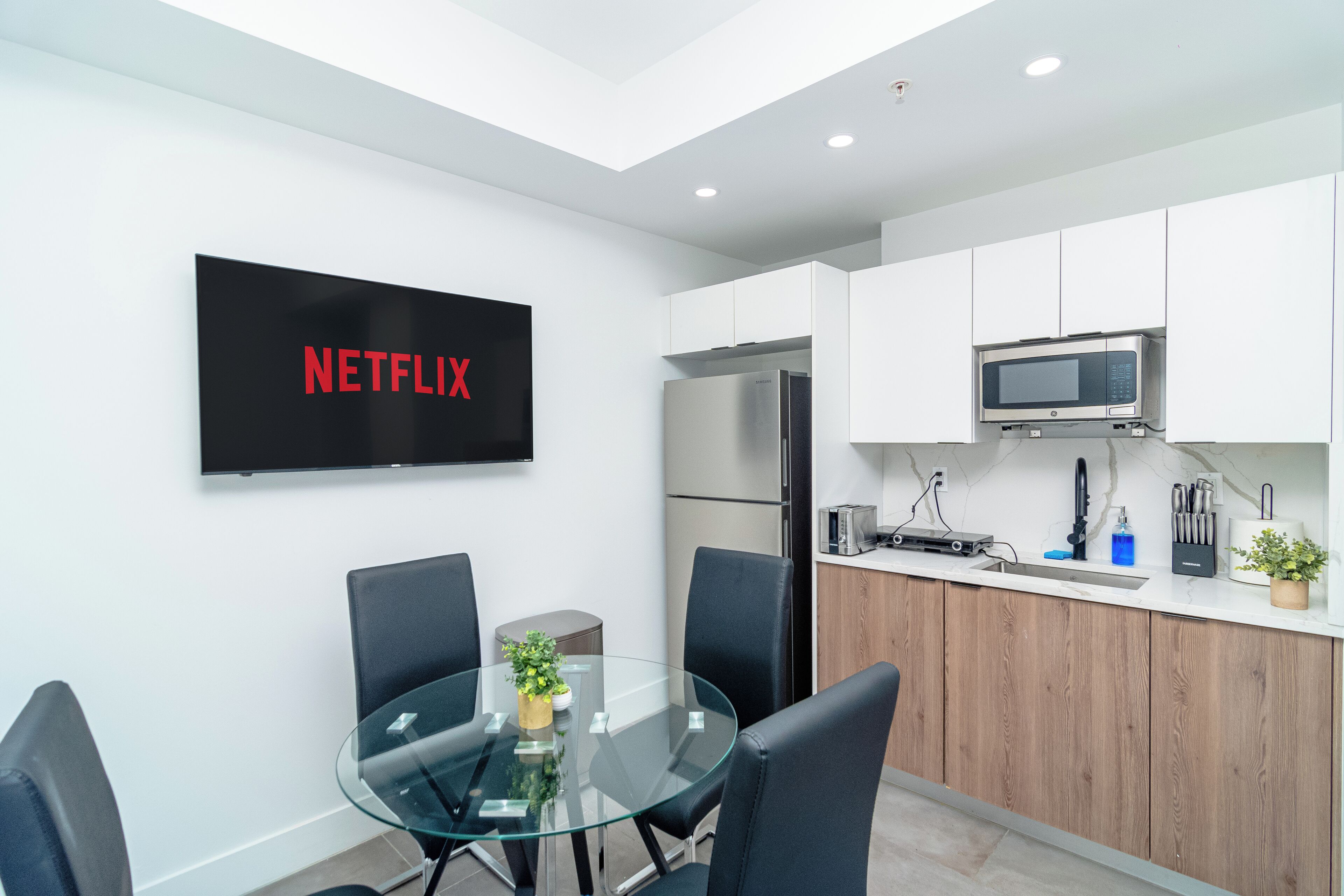 A modern kitchen with white cabinets, a stainless steel refrigerator, a microwave, and a glass dining table with chairs.