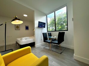 A modern room with a bed, a yellow armchair, a dining table with chairs, a TV mounted on the wall, and a large window with a view of trees. - Metro Park Hotel (Miami)