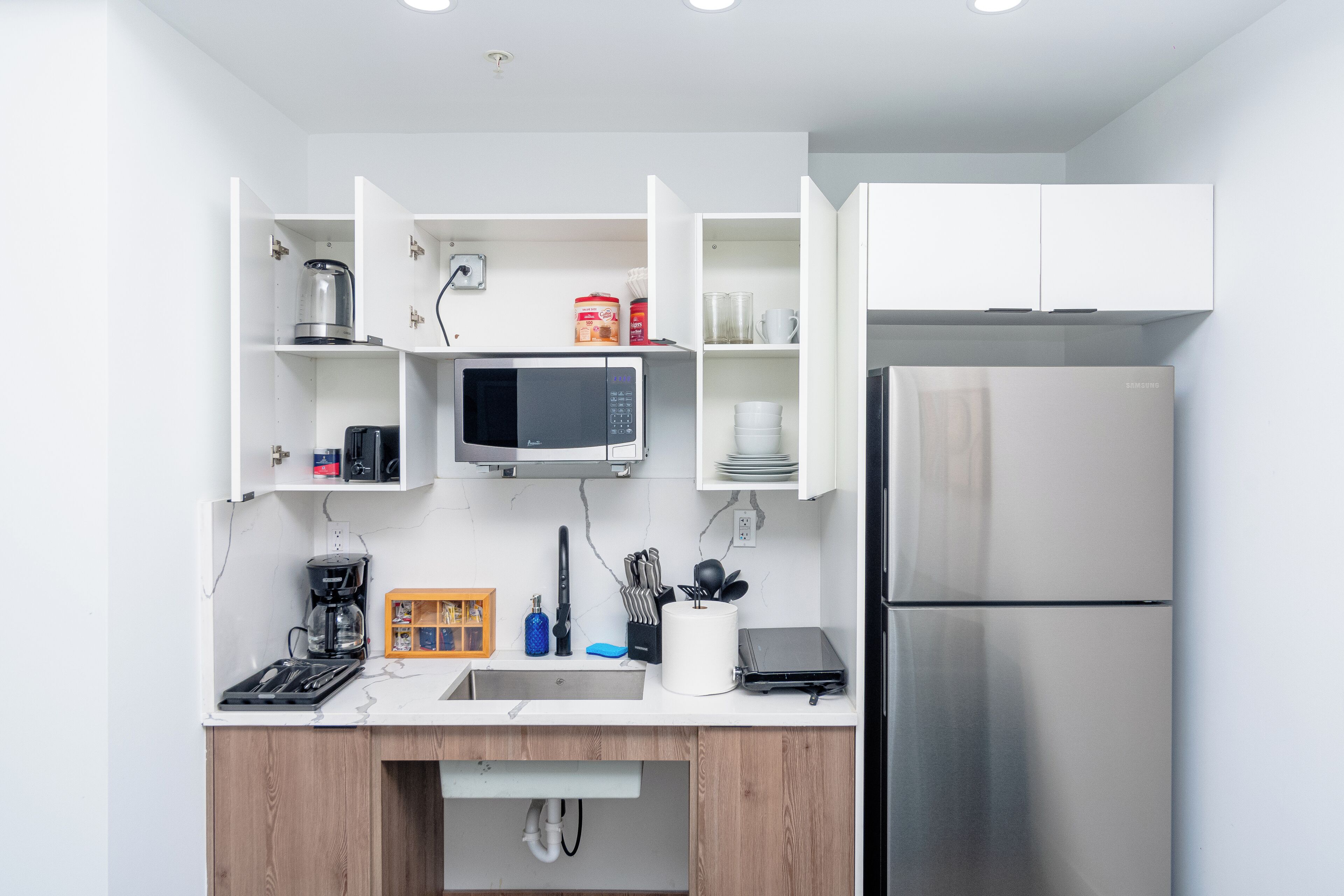 Una cocina moderna con muebles blancos, microondas integrado, refrigerador de acero inoxidable y cafetera.