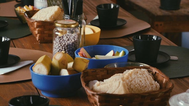 Una mesa de madera con diversos alimentos y recipientes, entre ellos, tazones de frutas, una cesta de pan y un tarro de frijoles.
