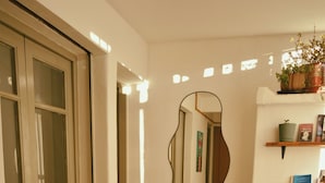 Una habitación con una puerta, un espejo, una planta en maceta, una estantería con libros y una cama.