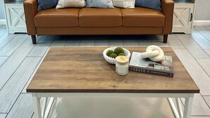 A modern living room with a brown leather sofa, two side tables with lamps, a coffee table with decorative items, and a large abstract painting on the wall. - The Bluebonnet Peak (Brownsville)