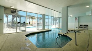 Una piscina interior con paredes de vidrio transparente, suelo de baldosas y vistas a palmeras y una zona junto a la piscina.