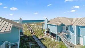 Ein direkt am Strand gelegenes Anwesen mit einer Holzstege, der zu einem Strand führt, mit einem blauen Gebäude mit Balkon und Treppe.