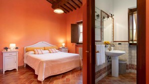 A bedroom with a bed, nightstands, a bedside lamp, a window, and a bathroom with a sink and mirror. - Villa with private pool and breathtaking views (Volterra)